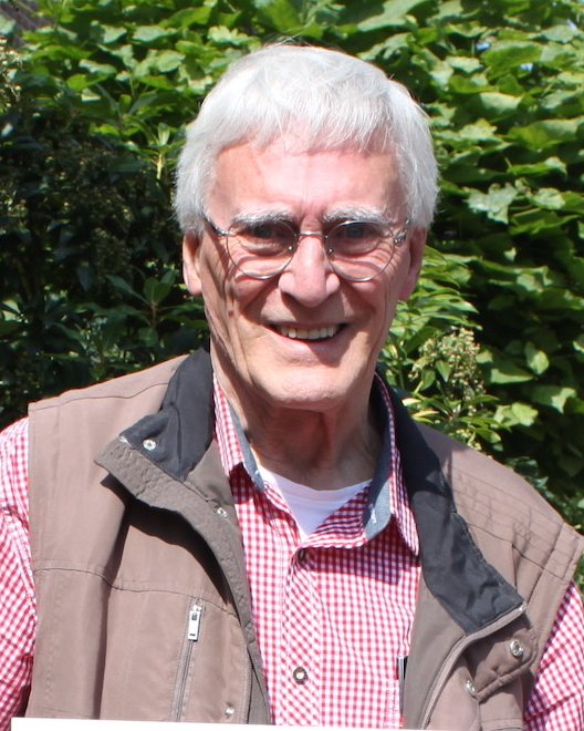 Portret van Leo Congert: een lachende man met grijs haar en een ronde bril, in een rood-wit geruit overhemd en een bruine bodywarmer, voor een groene haag.